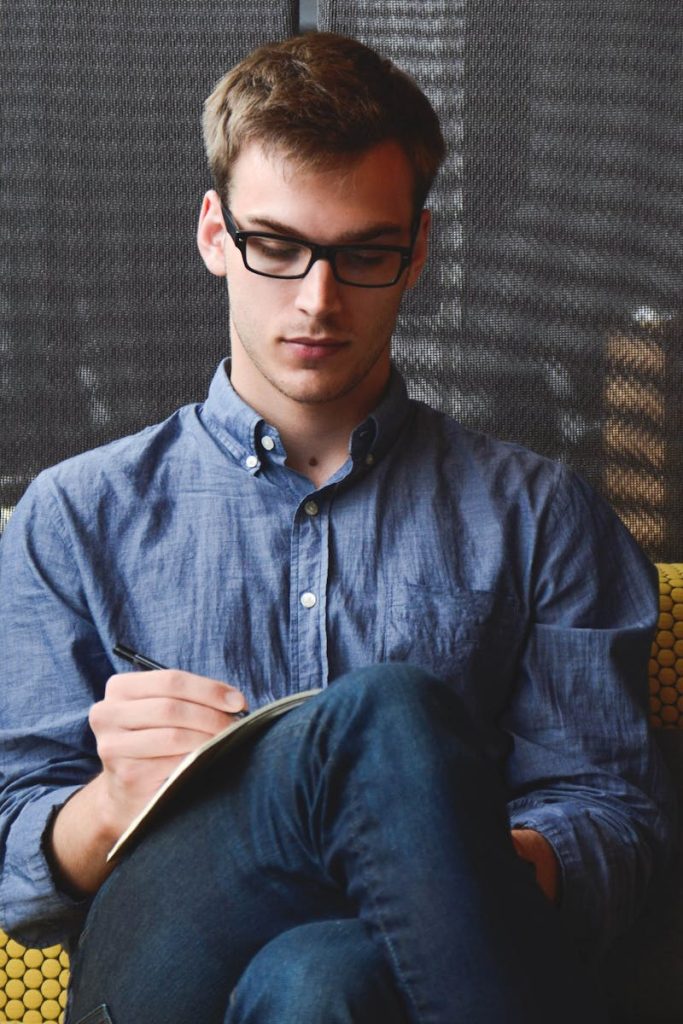 A young man in glasses writes in a notebook while sitting on a stylish couch indoors.